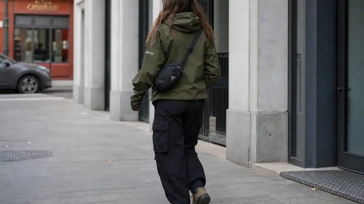Gorpcore celebrity street style — anonymous woman from behind in olive Gore-Tex shell jacket, black cargo pants, and brown hiking boots on urban sidewalk