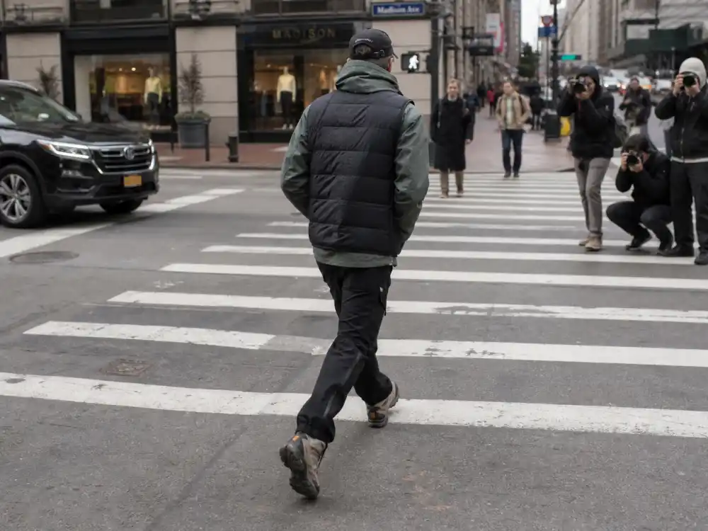 Anonymous figure from behind crossing city crosswalk in puffer vest, windbreaker, and hiking boots for gorpcore celebrity street style paparazzi moment