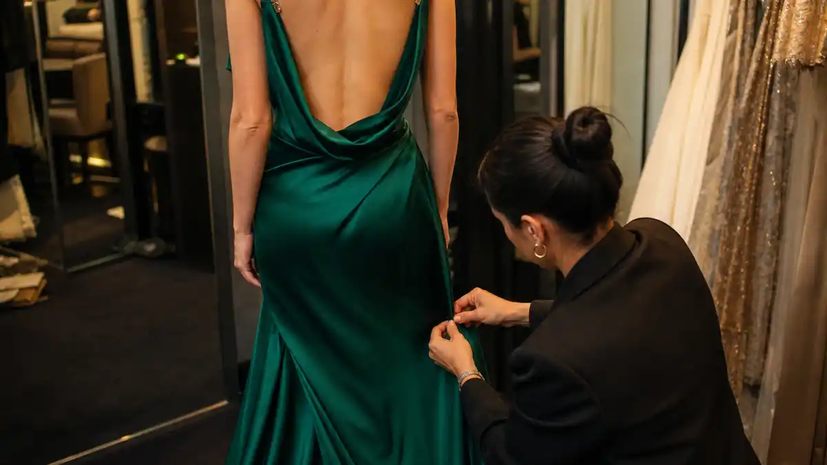 How celebrity stylists pick red carpet looks — stylist pinning emerald green satin gown hem on anonymous client from behind in luxury fitting room with garment racks