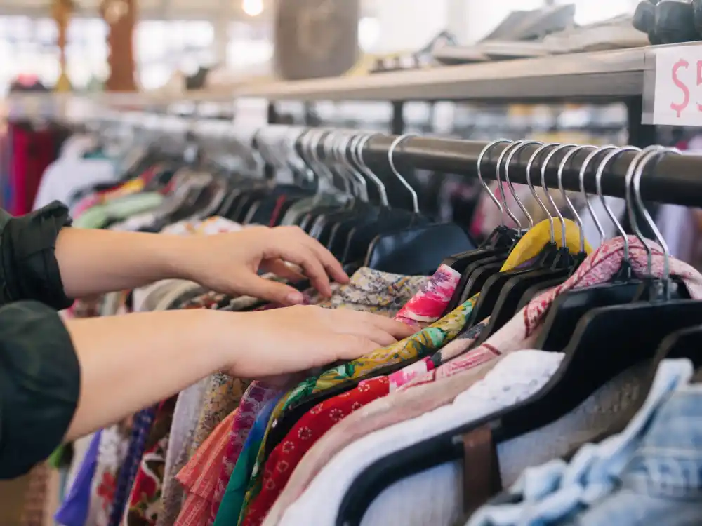 Hands browsing thrift store clothing racks central to how to dress like a celebrity on budget secondhand shopping strategy