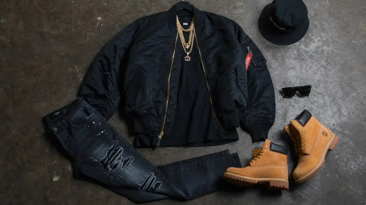 How to dress like AAP Rocky streetwear style — oversized black bomber jacket, layered gold chains, distressed slim jeans, wheat Timberland boots, bucket hat, and designer sunglasses flat lay