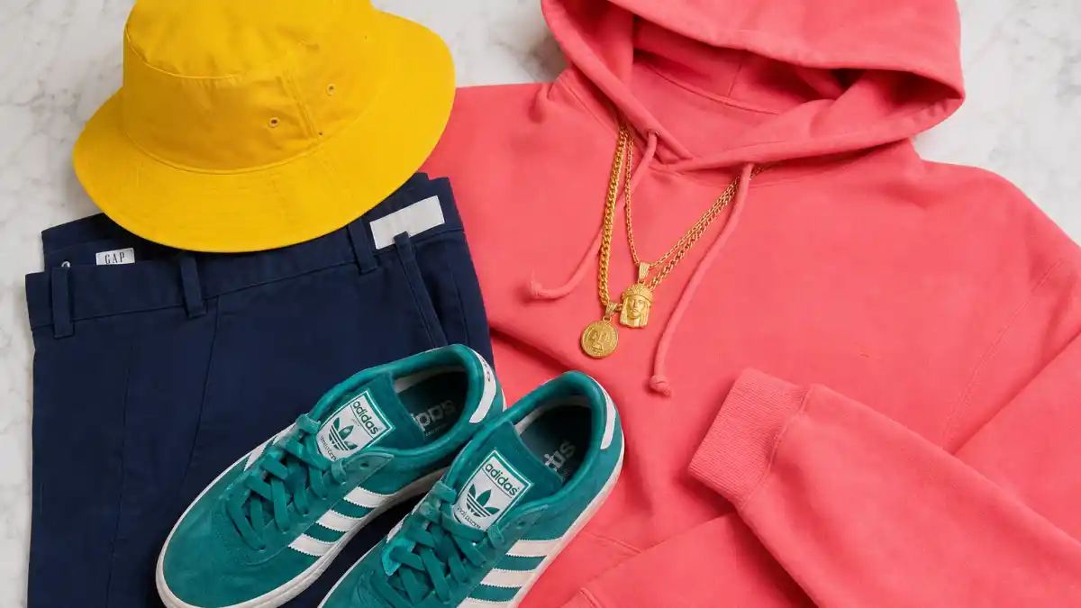 How to dress like Pharrell Williams colorful style — yellow bucket hat, coral pink hoodie, teal Adidas sneakers, navy chinos, and gold chains flat lay in bold streetwear color blocking