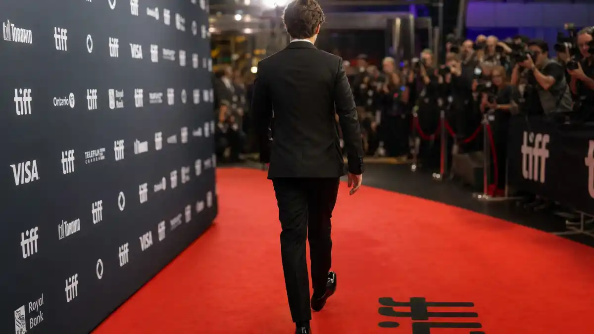 How to dress like Timothee Chalamet red carpet men — slim black tuxedo on premiere red carpet seen from behind with photographers and step-and-repeat backdrop