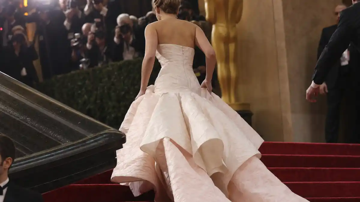 Jennifer Lawrence tripping Oscars dress moment — pale blush pink Dior couture ball gown from behind ascending Academy Awards stairs with paparazzi flashes