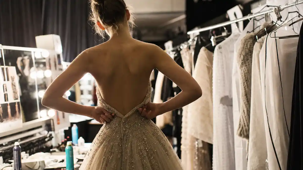 Kaia Gerber nepo baby modeling career — runway backstage from behind with designer gown and vanity mirrors editorial milestone without identifiable faces