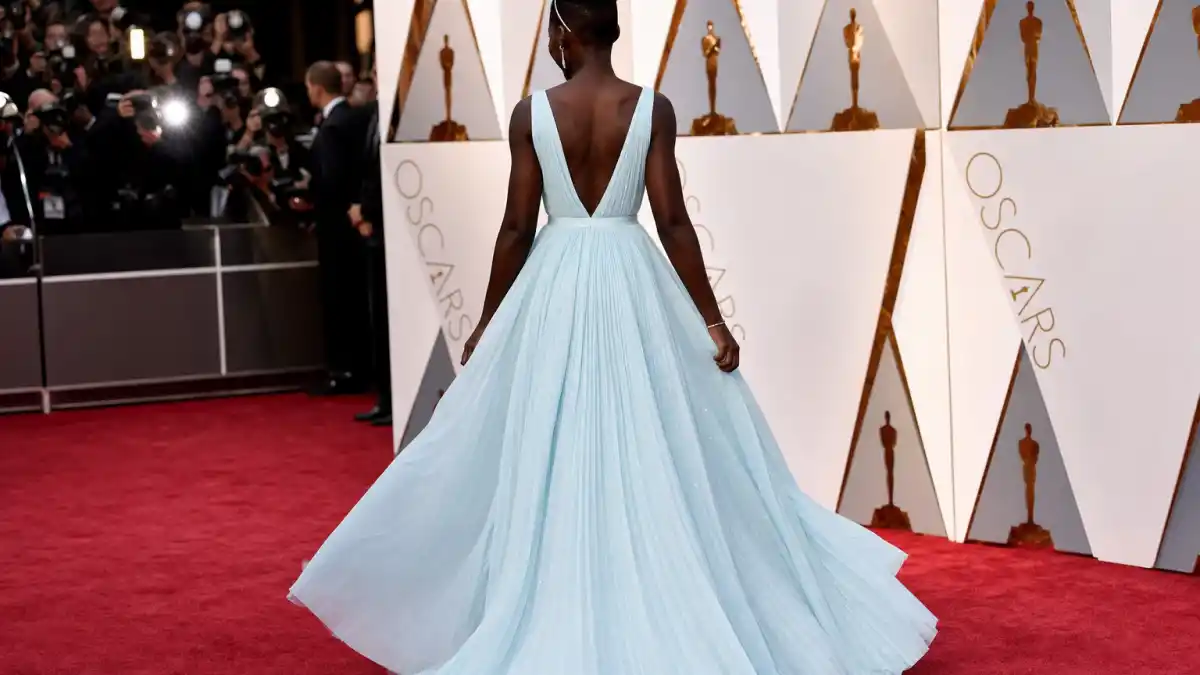 Lupita Nyongo blue Oscars gown — powder sky-blue pleated Prada silk dress photographed from behind on Academy Awards red carpet with Oscar statuette backdrop
