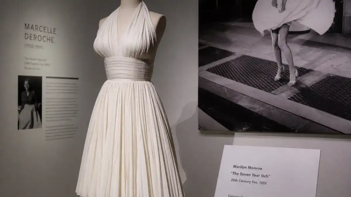 Marilyn Monroe white dress history — iconic white pleated halter cocktail dress displayed in fashion museum beside subway grate scene photograph from The Seven Year Itch