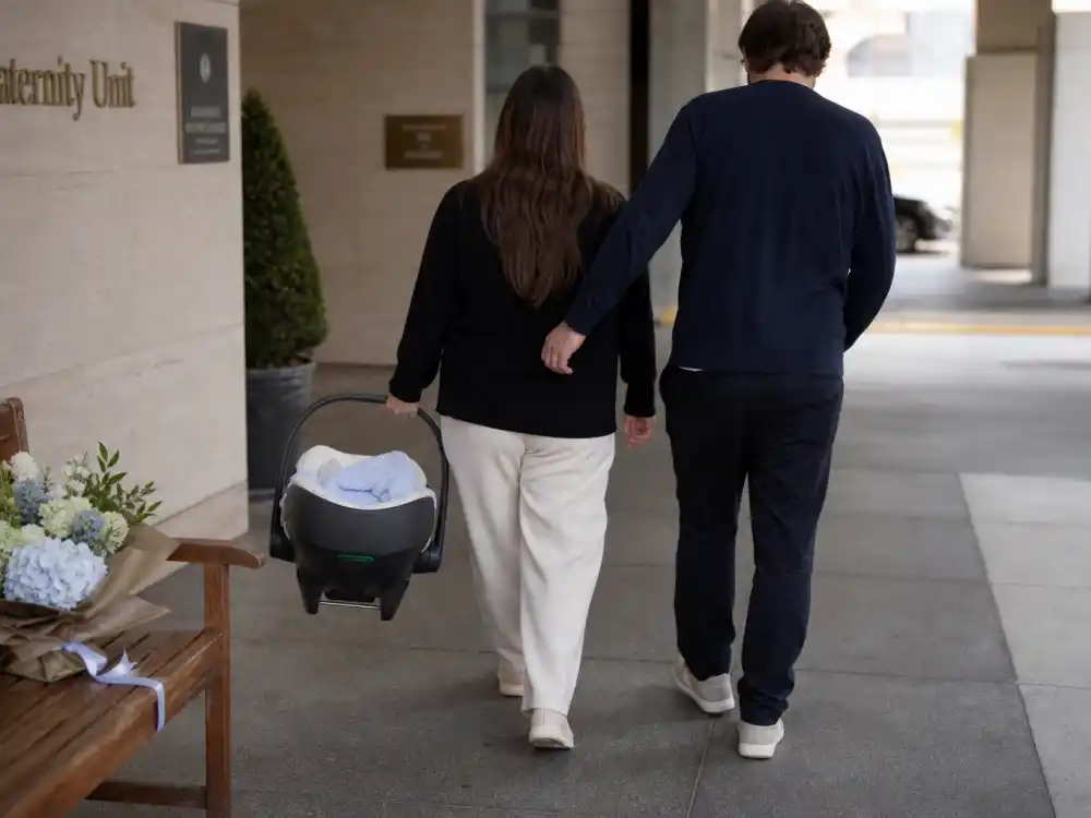 Hospital discharge scene for Meghan Trainor baby announcement 2026 — anonymous parents from behind with newborn bundle, no faces shown