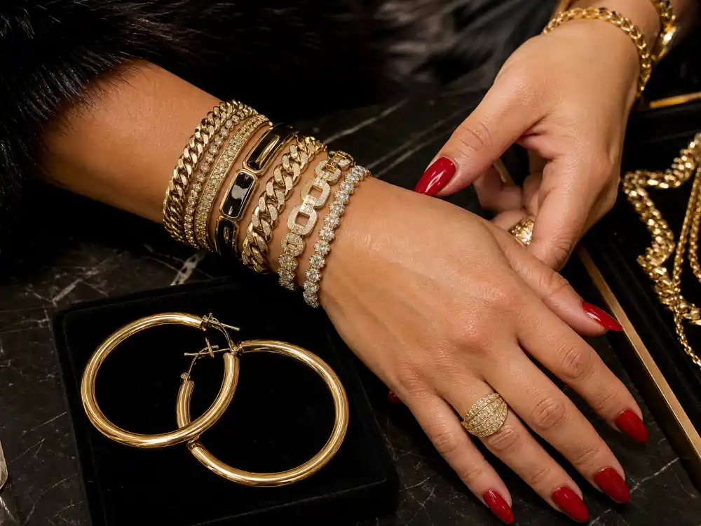 Stacked gold bracelets and large hoop earrings on velvet display with red manicured nails illustrating mob wife aesthetic celebrities jewelry layering rules