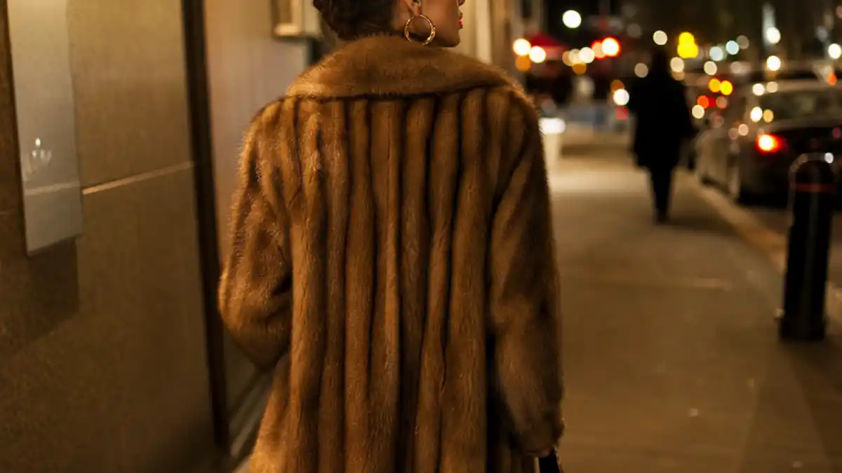 Mob wife aesthetic celebrities — anonymous woman from behind in full-length brown fur coat with gold hoop earrings and bold red lipstick on city sidewalk at night