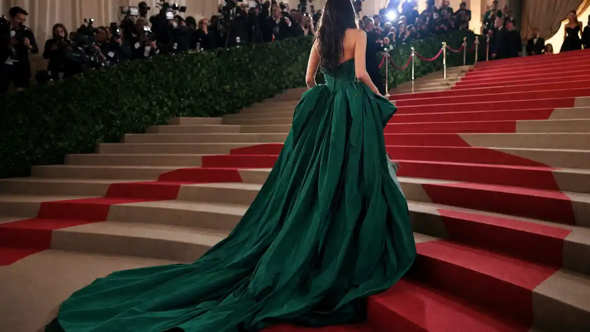 Most iconic Met Gala looks ever — anonymous woman from behind in emerald green couture ballgown ascending Metropolitan Museum grand staircase with paparazzi flash and red carpet runner
