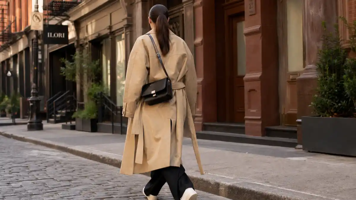 NYC celebrity street style trends — camel trench coat black wide-leg trousers white sneakers and crossbody bag on SoHo cobblestone sidewalk seen from behind