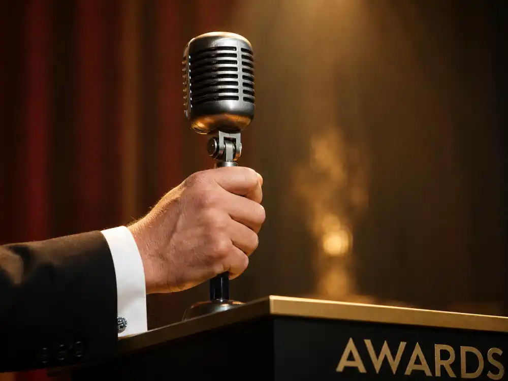A host hand grips the podium microphone under a spotlight — a defining image for the best Oscars hosts of all time commanding the Academy Awards stage
