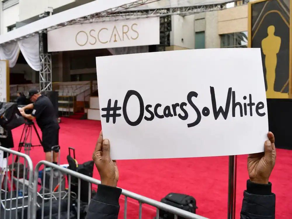 Anonymous hands holding OscarsSoWhite protest sign outside empty Oscar red carpet entrance capture the street-level anger behind OscarsSoWhite controversy explained