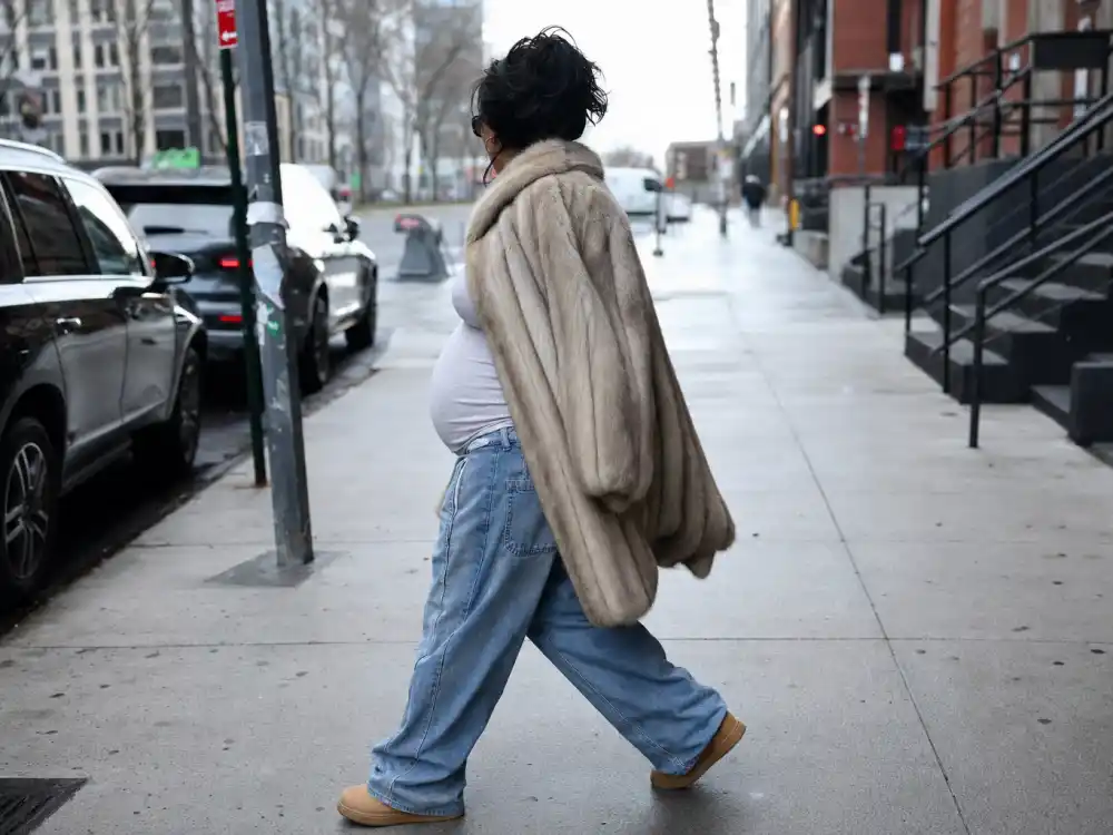 Anonymous pregnant woman in oversized vintage fur coat and baggy jeans on NYC sidewalk illustrating Rihanna pregnancy style evolution off-duty maternity streetwear