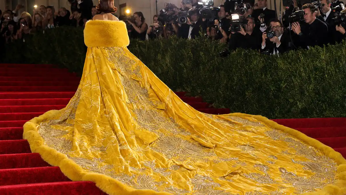 Rihanna yellow Met Gala cape dress — iconic golden-yellow Guo Pei couture gown with fur-trimmed cape and cathedral train on museum steps seen from behind