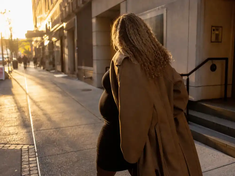 Anonymous pregnant woman in camel coat and fitted dress on NYC sidewalk for Serena Williams maternity fashion looks off-duty pregnancy street style