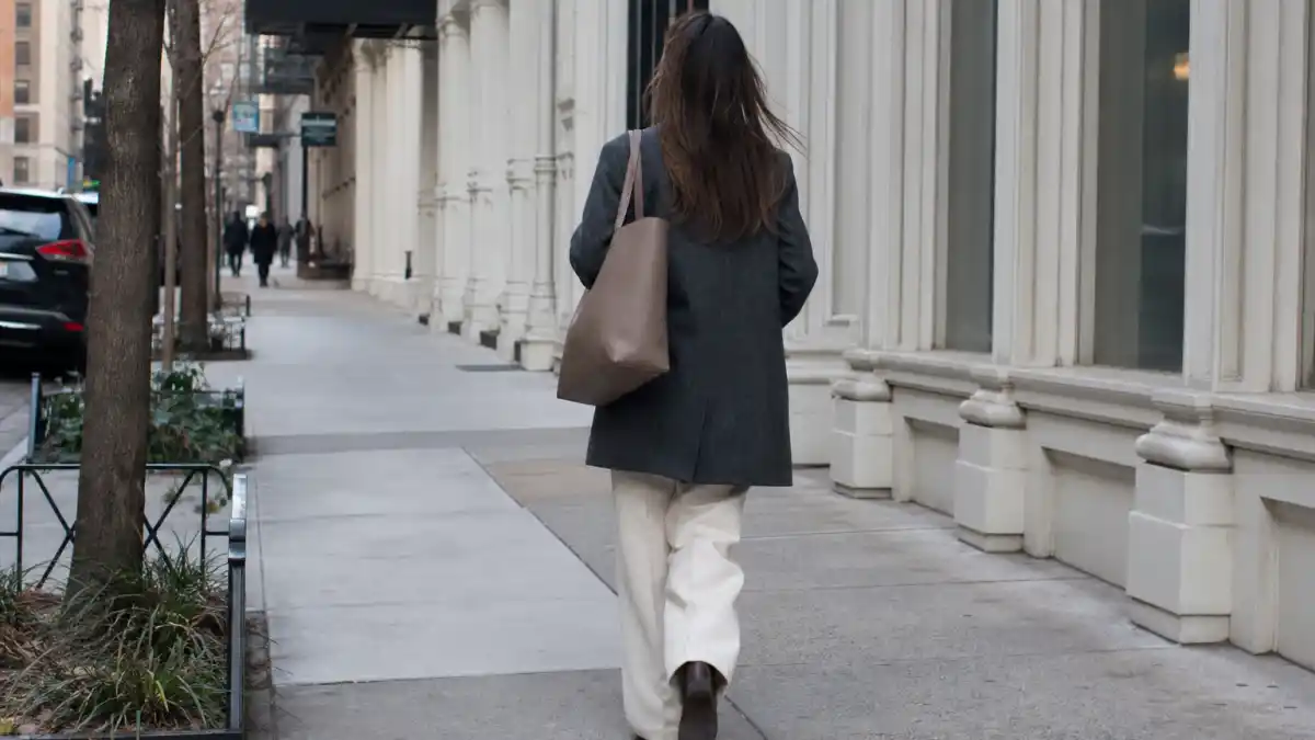 Stealth wealth celebrity fashion — anonymous woman from behind in charcoal wool coat and cream wide-leg trousers on upscale city sidewalk with unstructured taupe leather tote and no visible logos