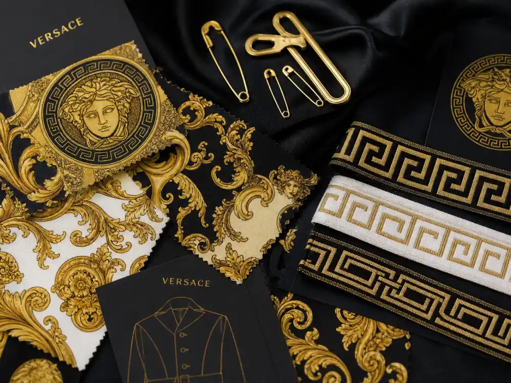Baroque print fabric swatches gold safety pins and Greek key trim flat lay capturing print language behind legendary Versace celebrity red carpet moments worldwide