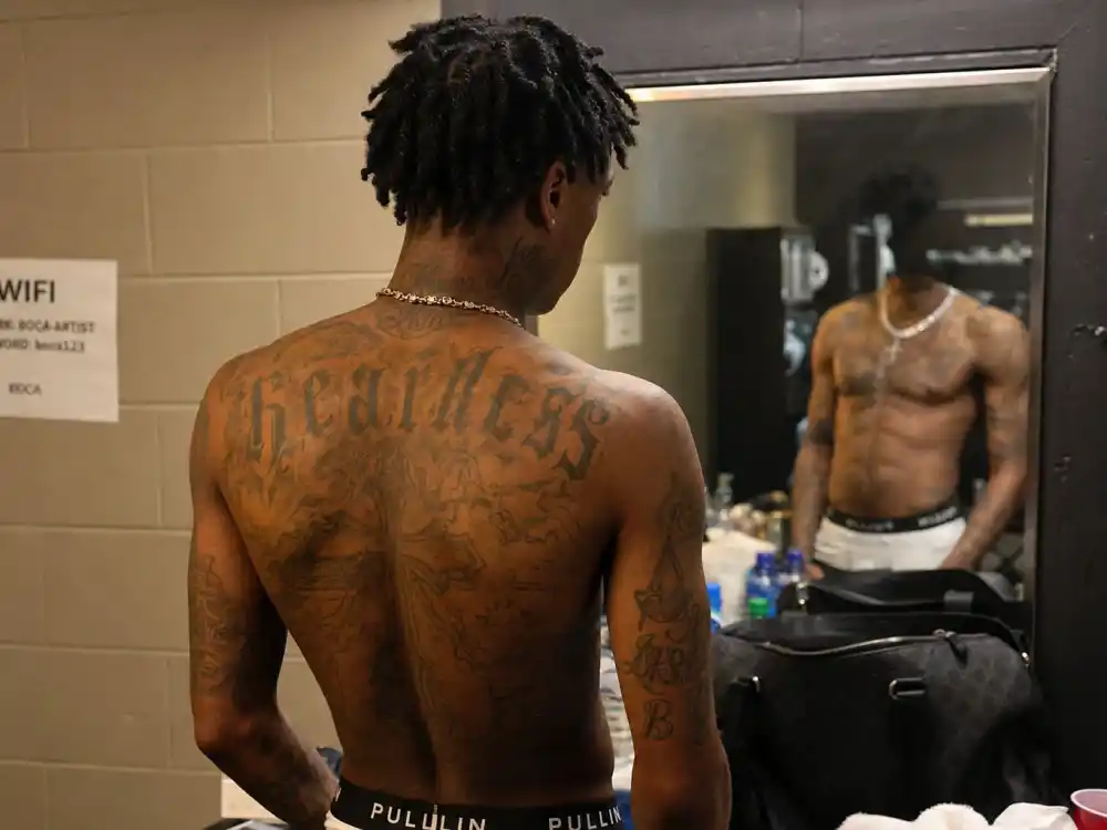 21 Savage tattoos and their meanings — chest cross and script tattoos visible over-shoulder on shirtless rapper backstage