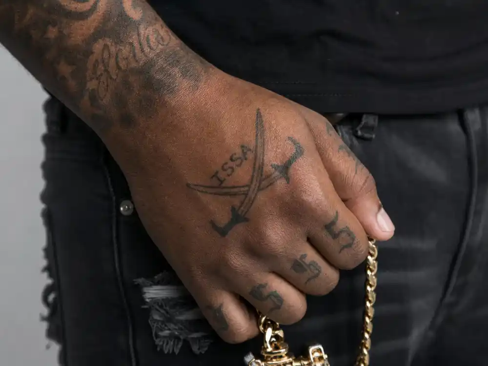 21 Savage tattoos and their meanings — crossed swords ISSA hand tattoo and finger ink on rapper hand holding chain