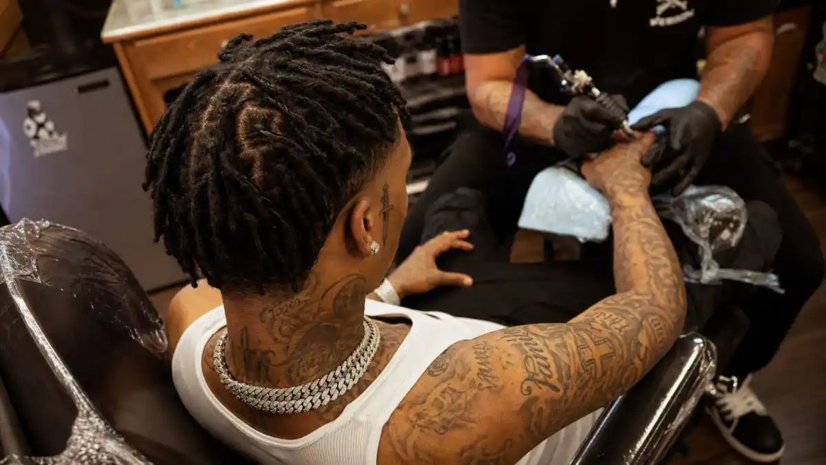 21 Savage tattoos and their meanings — Atlanta rapper seen from behind in tattoo studio with signature forehead dagger tattoo and full arm sleeves visible
