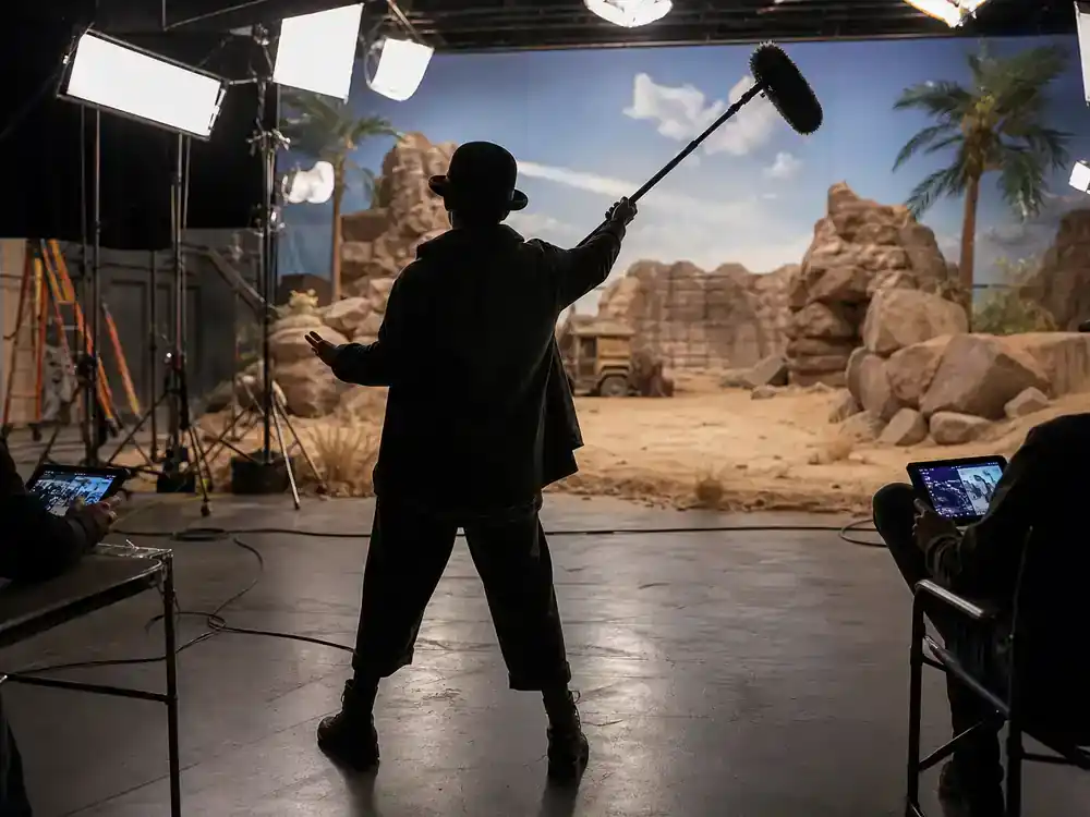A Minecraft Movie cast and cameos guide Steve — comedic actor silhouette rehearsing on soundstage with boom mic and crew tablets, no identifiable face