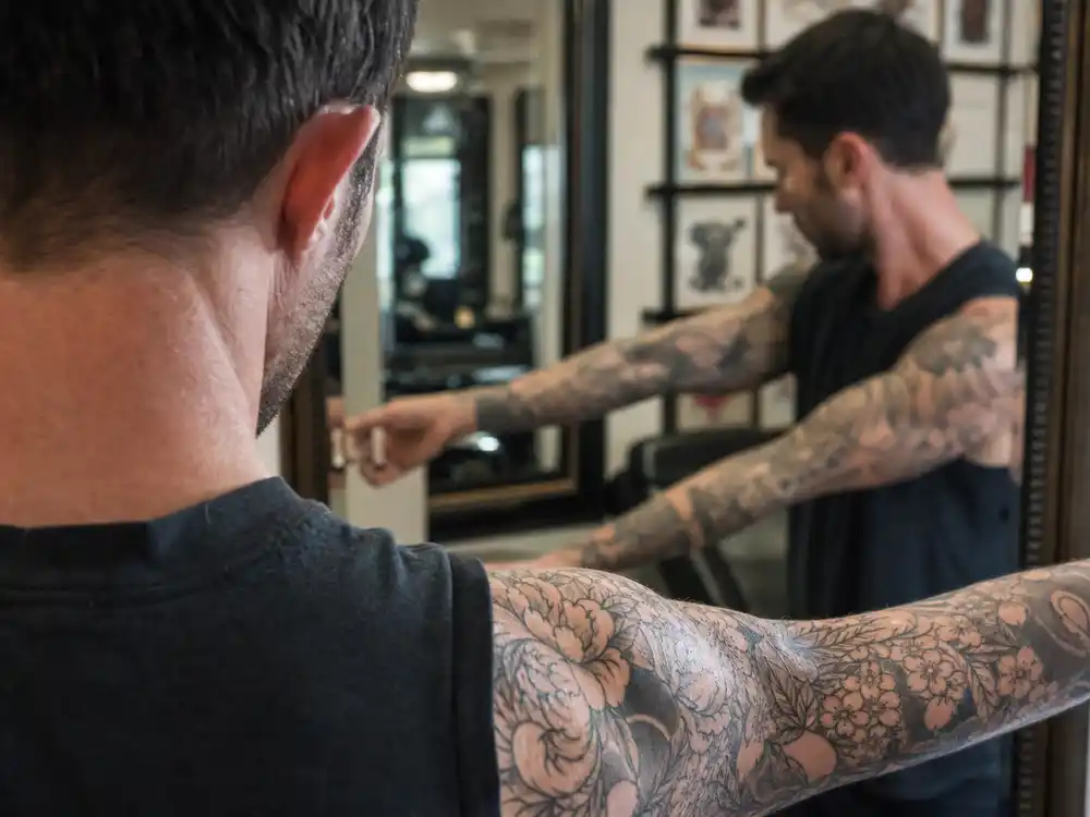 Adam Levine tattoo collection guide cherry blossom sleeve — close-up of floral black grey forearm tattoos on lean singer in studio mirror