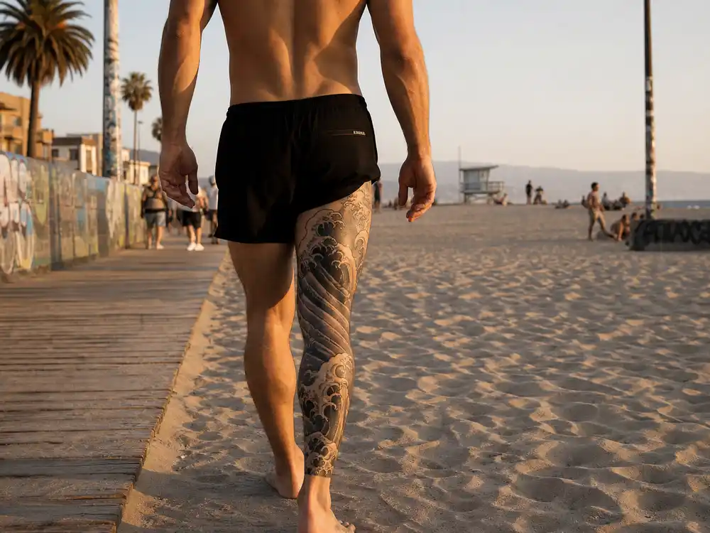 Adam Levine tattoo collection guide leg waves — back view at California beach showing Japanese-style black grey wave tattoo wrapping left leg
