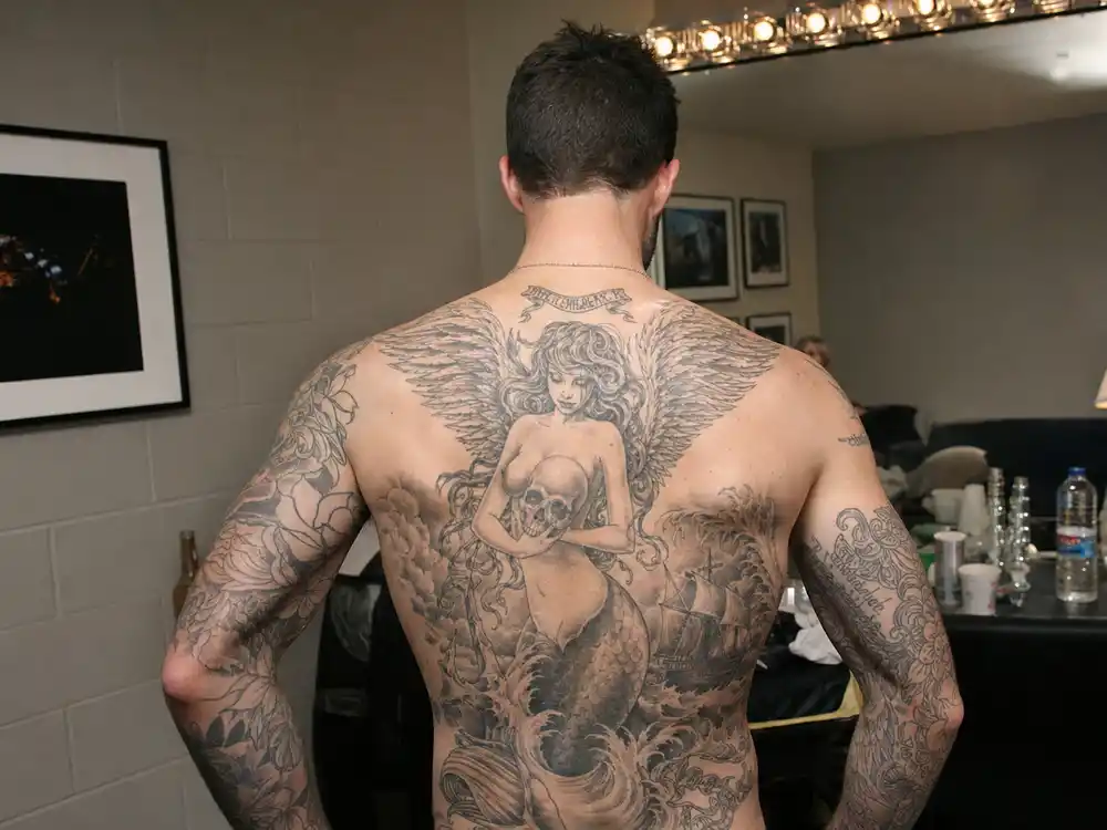 Adam Levine tattoo collection guide mermaid back mural — singer from behind showing siren wings skull waves and lost ship back piece in green room