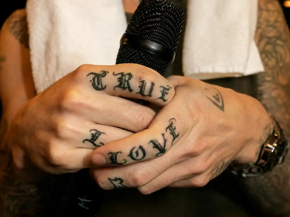 Adam Levine tattoo collection guide True Love fingers — close-up of script finger tattoos holding microphone backstage at concert