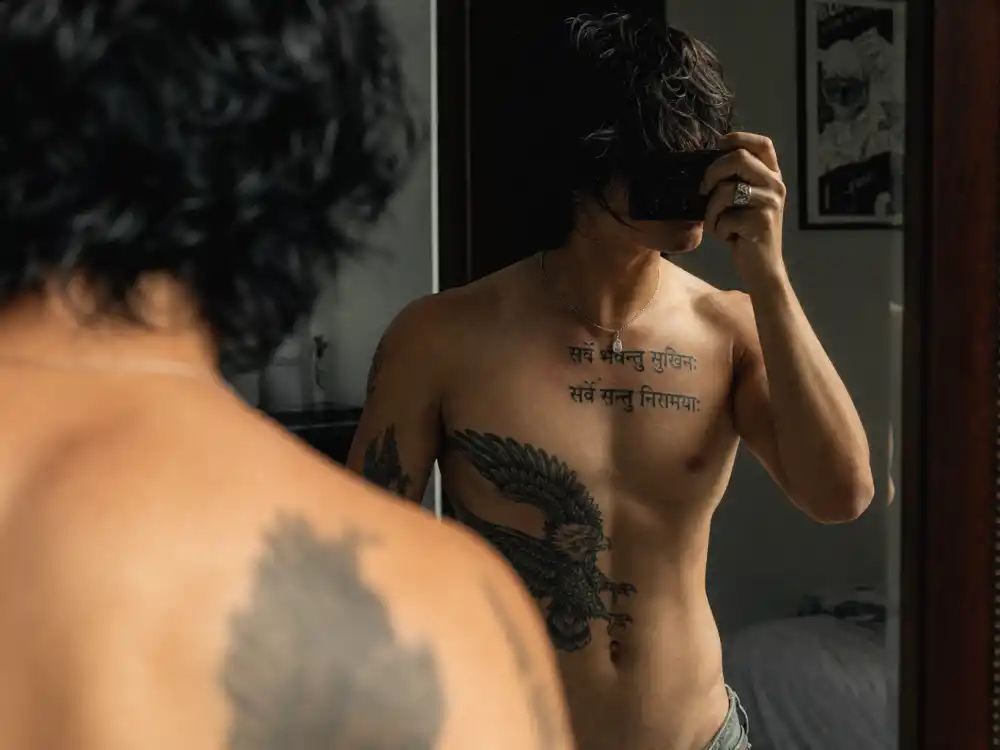 Adam Levine tattoos and their meanings chest piece — Sanskrit tapas script and eagle rib ink over-shoulder mirror angle