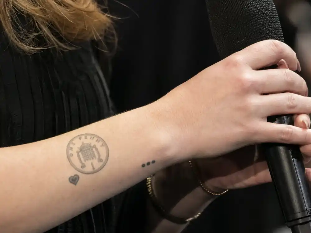 Adele tattoos and their meanings One Penny — minimalist coin tattoo reading ONE PENNY with heart on left wrist close-up at tattoo studio
