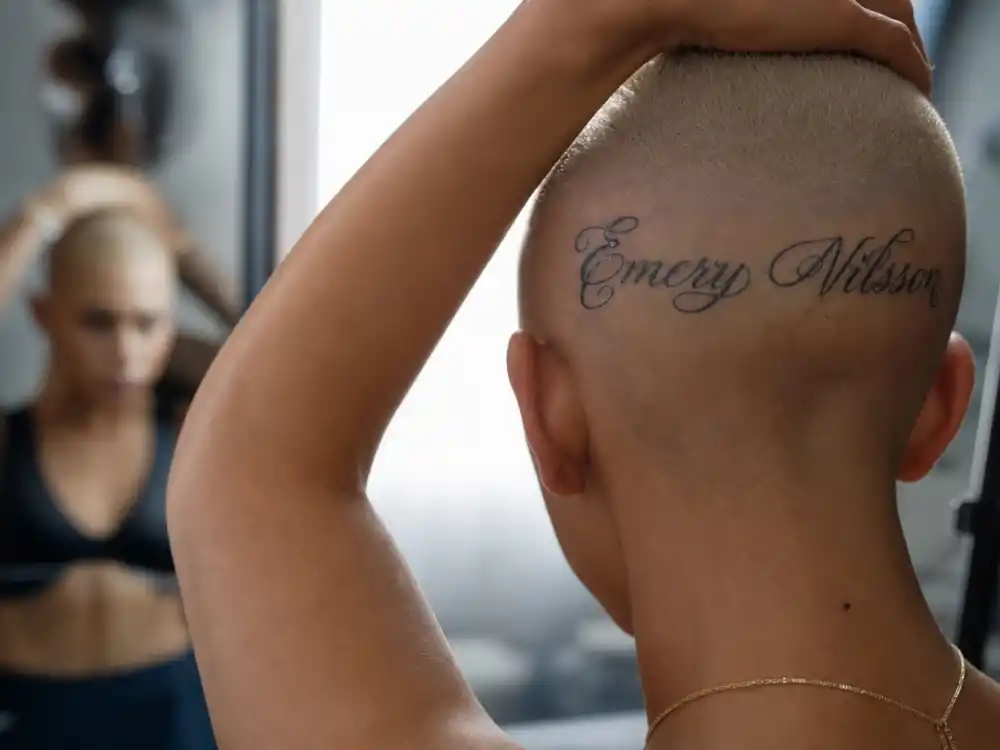 Amber Rose tattoos and their meanings — close-up of Bash Slash forehead script tattoo on shaved blonde head from over-shoulder angle