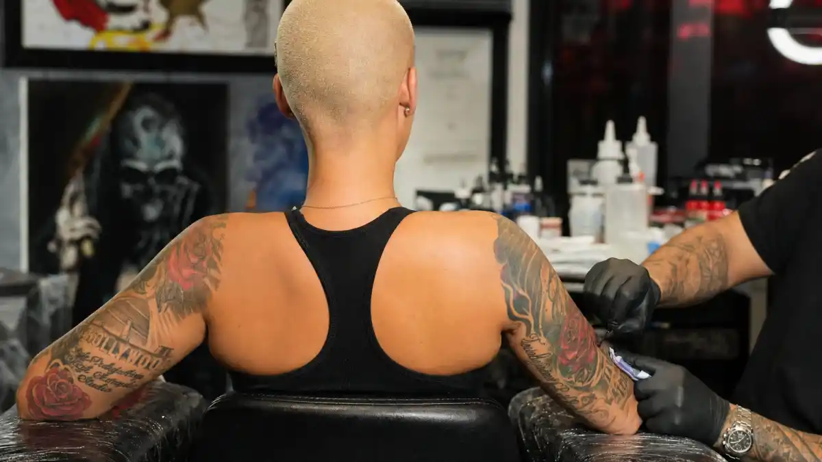Amber Rose tattoos and their meanings — model seen from behind in tattoo studio with rose sleeve ink and Slash portrait forearm visible