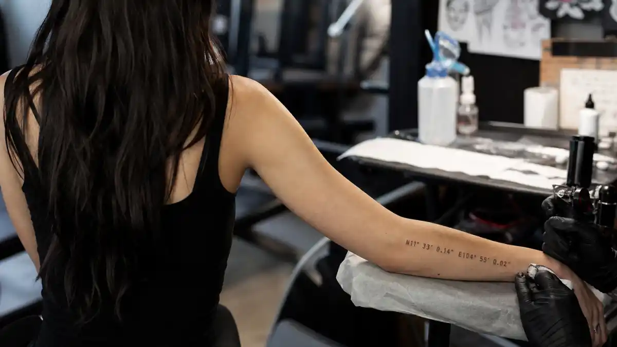 Angelina Jolie tattoos and their meanings — actress seen from behind in tattoo studio with geographic coordinate forearm ink and lower back tiger tattoo visible