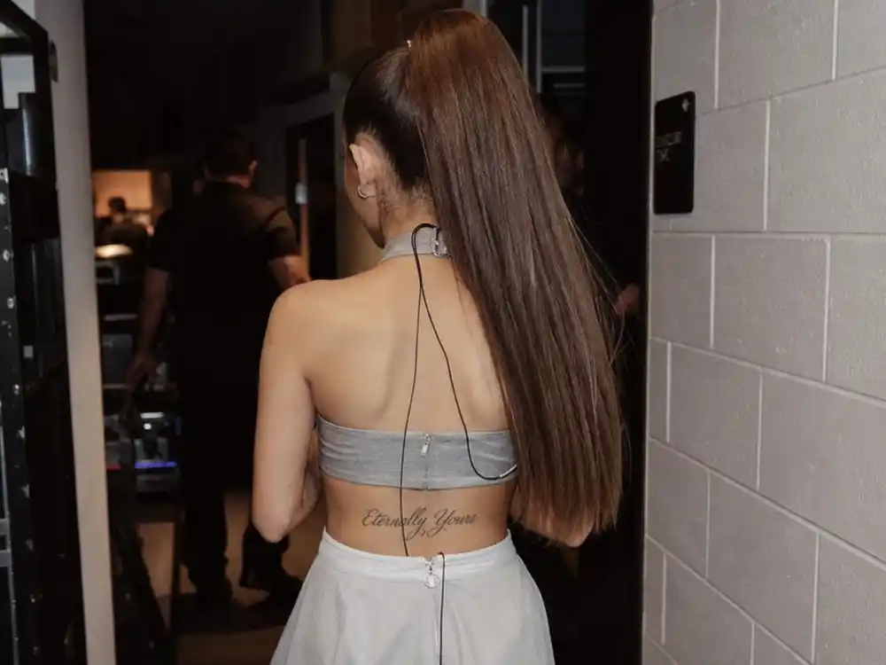 Ariana Grande tattoos and their meanings — Eternally Yours script tattoo on lower back seen from behind in backstage corridor