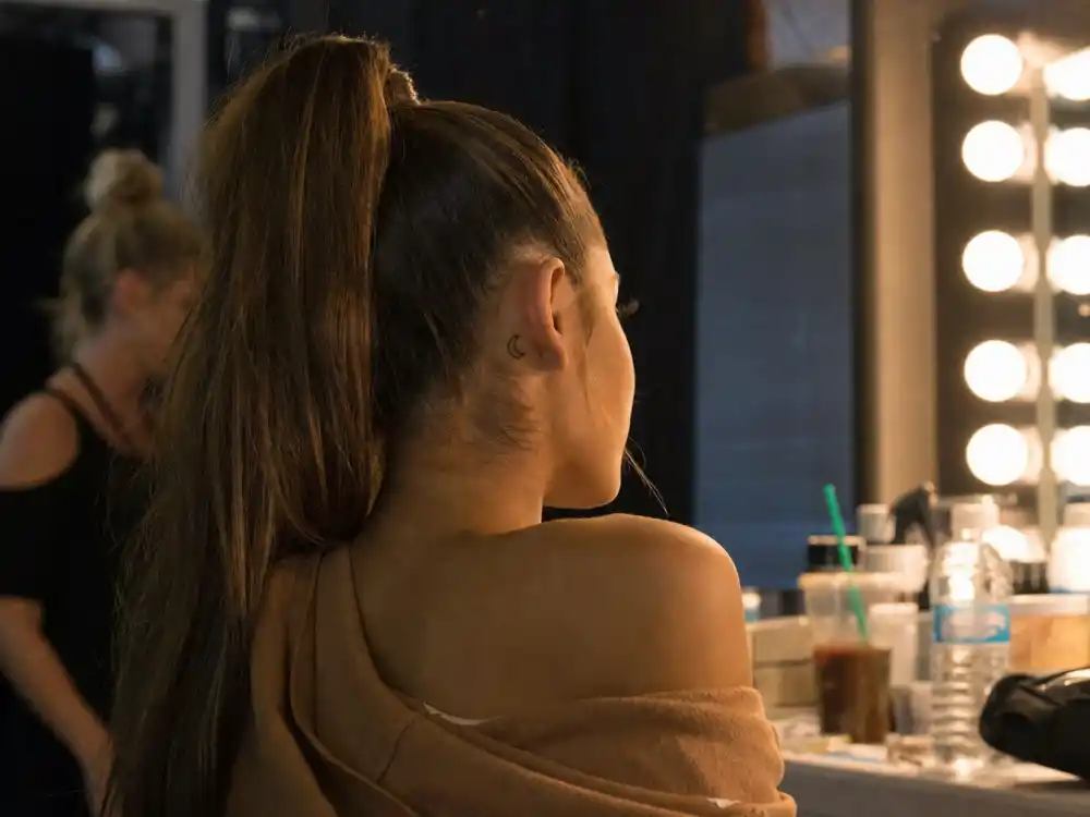 Ariana Grande tattoos and their meanings — crescent moon tattoo behind ear photographed in dressing room mirror over-shoulder angle