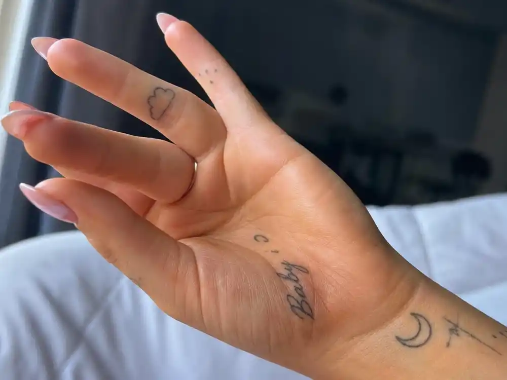 Ariana Grande tattoos and their meanings — close-up of cloud finger tattoo, heart shapes, and Baby I palm script in fine-line ink