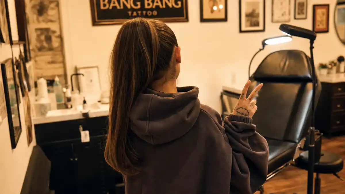 Ariana Grande tattoos and their meanings — pop star seen from behind in tattoo studio with delicate hand tattoos, cloud finger ink, and high ponytail visible