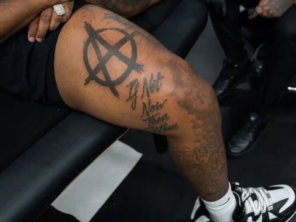 ASAP Rocky tattoos and their meanings — anarchy symbol and If Not Now Then When thigh script on rapper leg in studio