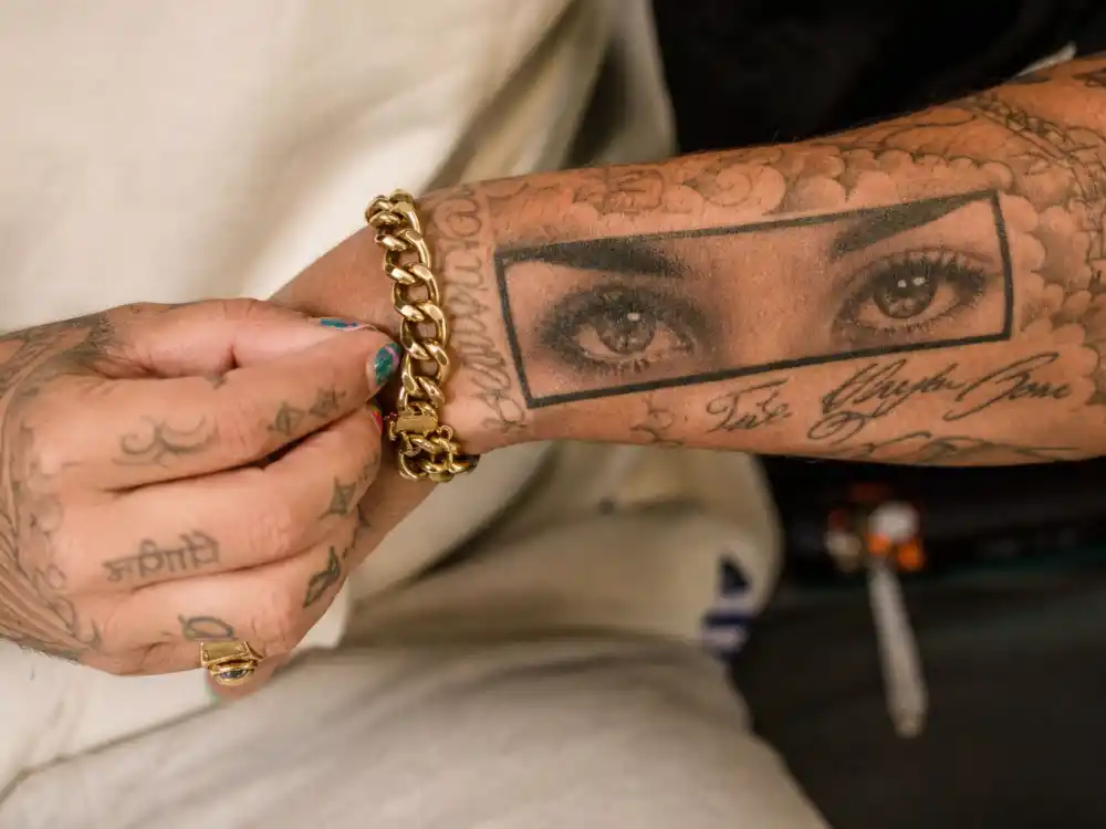 Bad Bunny tattoos and their meanings — close-up of realistic pair of eyes forearm tattoo and finger script on hand with colorful nail polish