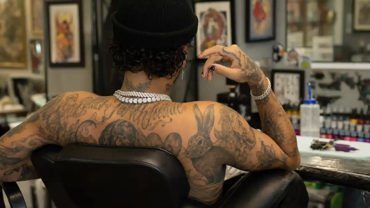 Bad Bunny tattoos and their meanings — Puerto Rican reggaeton artist seen from behind in tattoo studio with curly hair, colorful nails, and extensive arm and neck ink visible