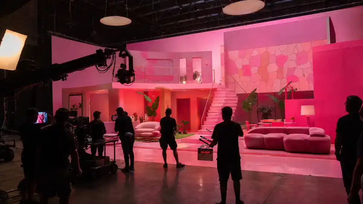 Barbie movie filming locations and set design — massive bright pink film soundstage with camera crane clapperboard and crew silhouettes on blockbuster studio set