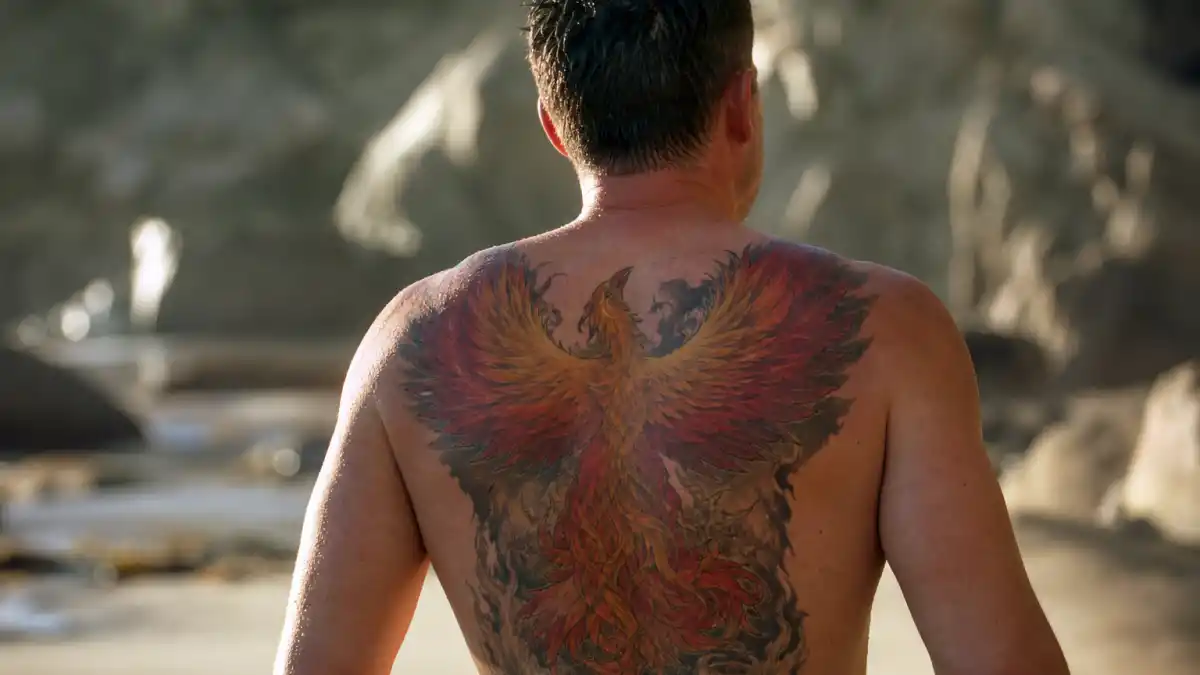 Ben Affleck tattoos and their meanings — actor seen from behind on Malibu beach with massive colorful phoenix back tattoo visible