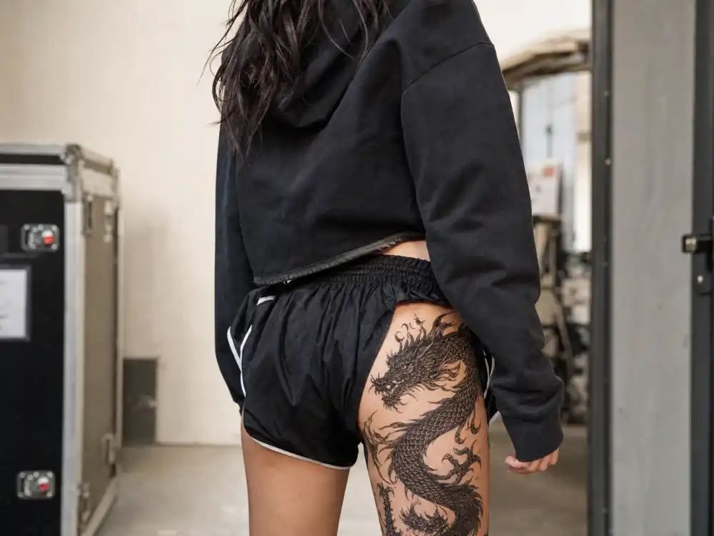 Billie Eilish tattoos and their meanings dragon — large black dragon tattoo on right thigh and hip seen from behind in backstage corridor