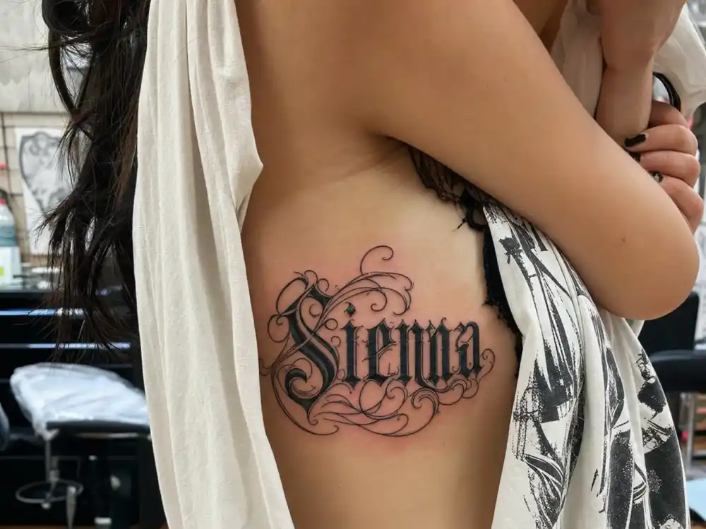 Billie Eilish tattoos and their meanings Eilish — ornate gothic script middle name tattoo on ribcage close-up at tattoo studio