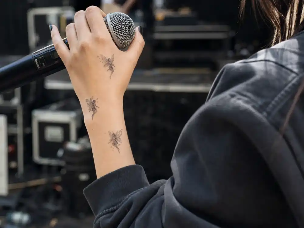 Billie Eilish tattoos and their meanings fairies — three delicate fairy tattoos on left hand and wrist close-up backstage