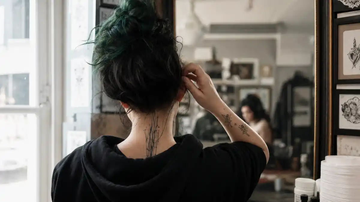 Billie Eilish tattoos and their meanings — pop singer from behind at tattoo studio with fairy wrist ink and spine tattoo at nape visible