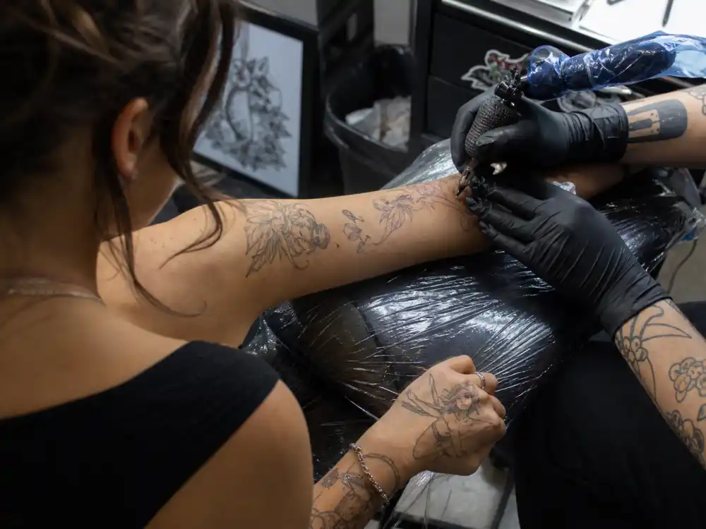 Billie Eilish tattoos and their meanings studio session — tattoo artist gloved hands applying ink on forearm over-shoulder with fairy hand tattoos visible